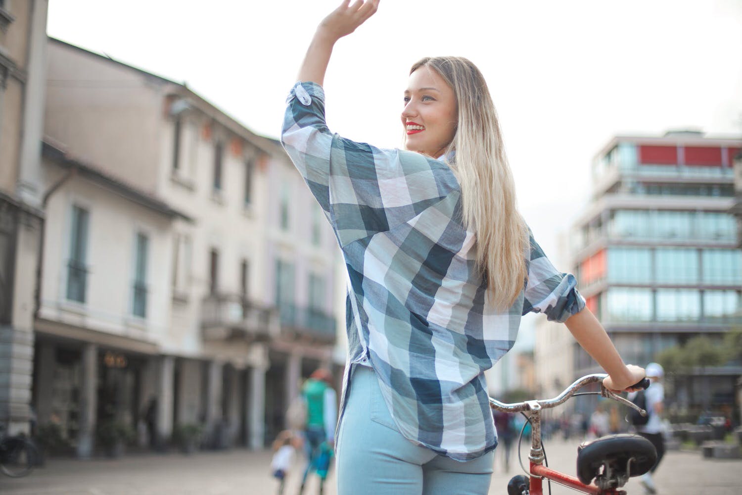 back view photo of smiling woman in checkered shirt and blue denim jeans standing with a bicycle waving